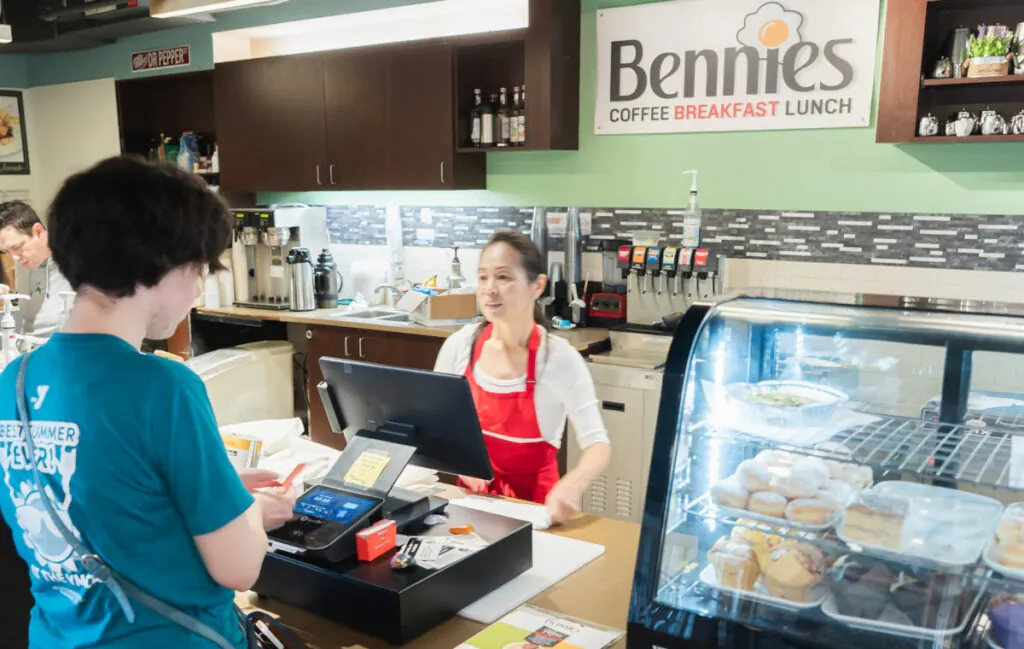 A customer at a checkout counter with a card reader and register, being served by a woman in a red apron at a cafe with a sign reading Bennies Coffee Breakfast Lunch, with a pastry display case full of muffins and desserts in the foreground.
