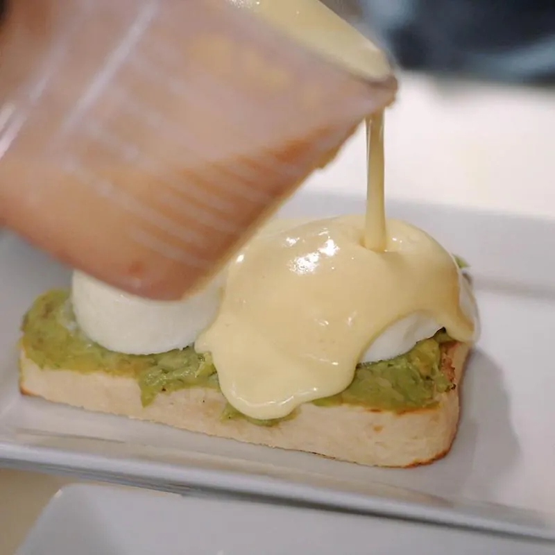 A hand pouring yellow hollandaise sauce from a plastic measuring cup over a poached egg on mashed avocado toast, plated on a white square dish.