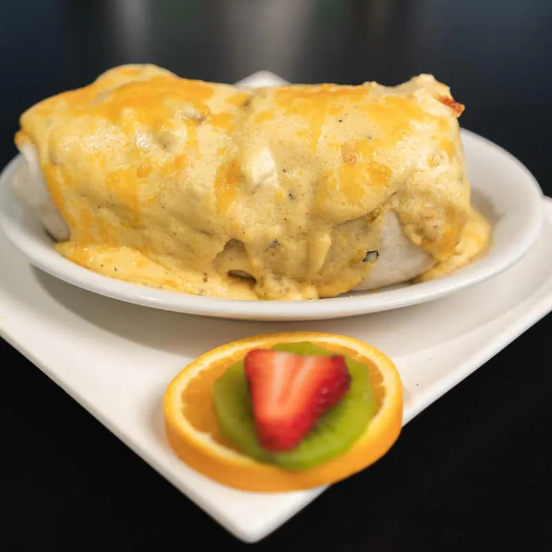A large burrito smothered in melted cheese sauce on a round white plate, garnished with an orange slice topped with kiwi and strawberry, set on a black table.