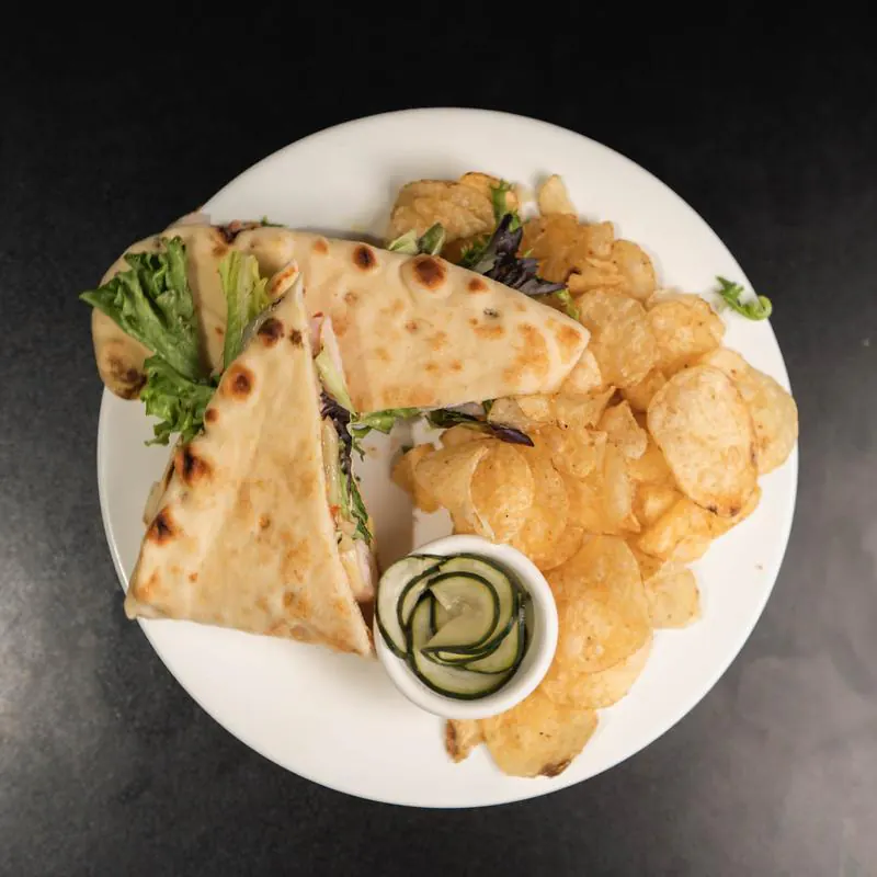 A white plate with a flatbread sandwich cut into triangles and filled with lettuce and meat, served alongside a pile of potato chips and a small cup of pickled cucumber slices.