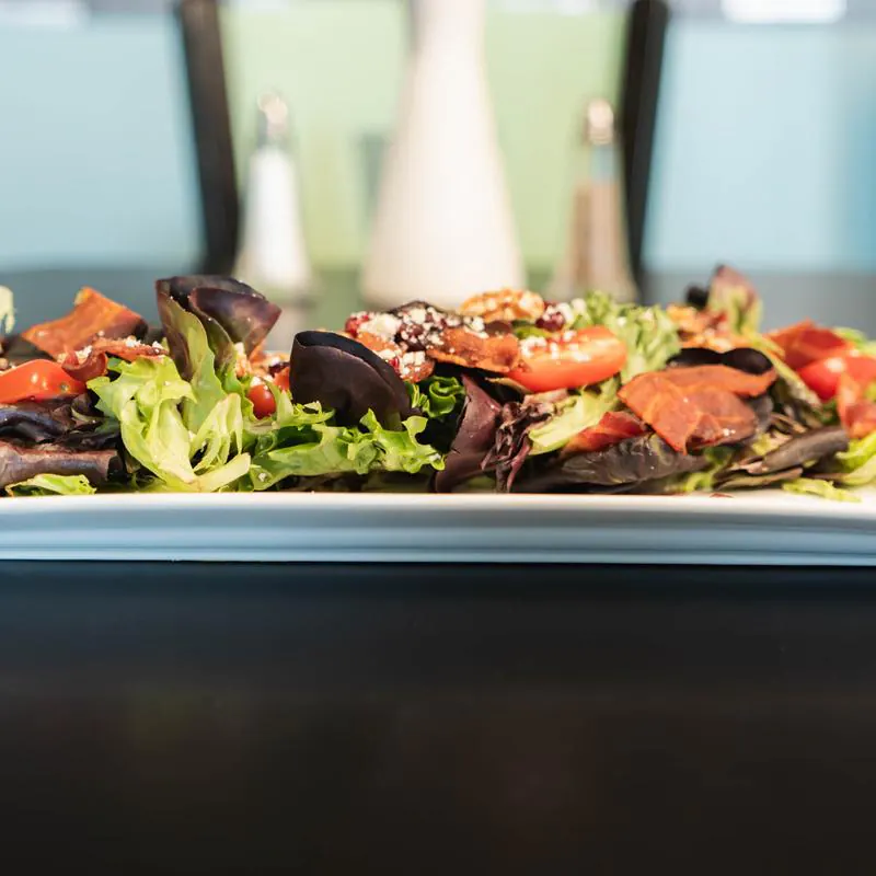 A long white plate holding a salad of mixed greens, cherry tomatoes, crumbled cheese, nuts, and crispy bacon pieces, with salt and pepper shakers visible in the blurred background.