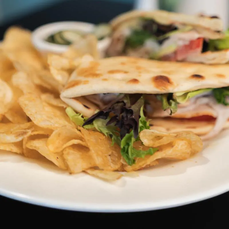 A white plate with a flatbread wrap sandwich filled with lettuce, tomato, and meat, cut in half and served with a pile of potato chips and a small cup of pickle slices.