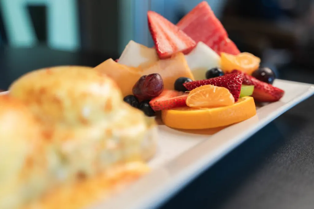 A rectangular white plate holding sliced fresh fruit including strawberries, watermelon, cantaloupe, orange slices, grapes, and blueberries, with a blurred pastry in the foreground.
