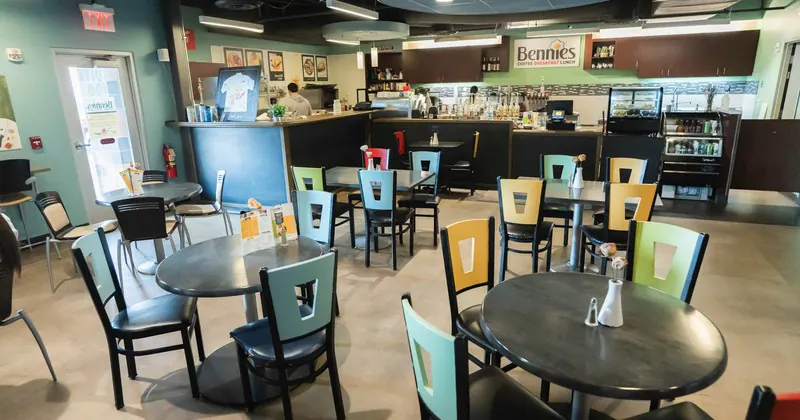 Interior of a cafe named Bennies with a service counter, colorful chairs with cutout backs around round tables, and a beverage cooler along the wall.