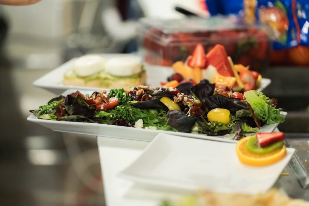A long white platter of mixed greens salad with cherry tomatoes, nuts, cranberries and cheese crumbles, set on a buffet table with fruit skewers, sliced fruit and a bread dish visible in the background.