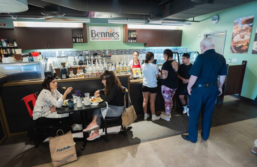 Interior of a cafe called Bennies Coffee Breakfast Lunch, with two women seated eating at a small table, several customers lined up at the counter, a staff member behind the counter, and shelves of syrup bottles and dispensers along the wall.