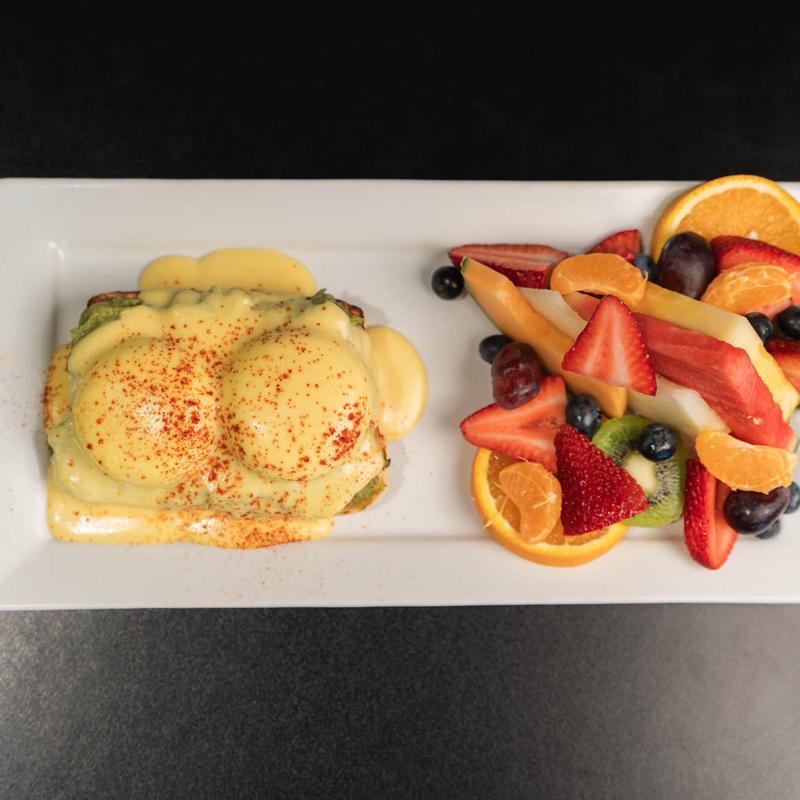 A rectangular white plate with eggs Benedict topped with hollandaise sauce and paprika, served alongside a mixed fruit salad of strawberries, blueberries, kiwi, melon, grapes, and citrus slices.